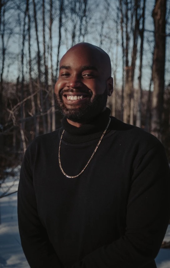 Roy AT Williams, smiling, in a black turtleneck and gold chain outdoors in a winter forest